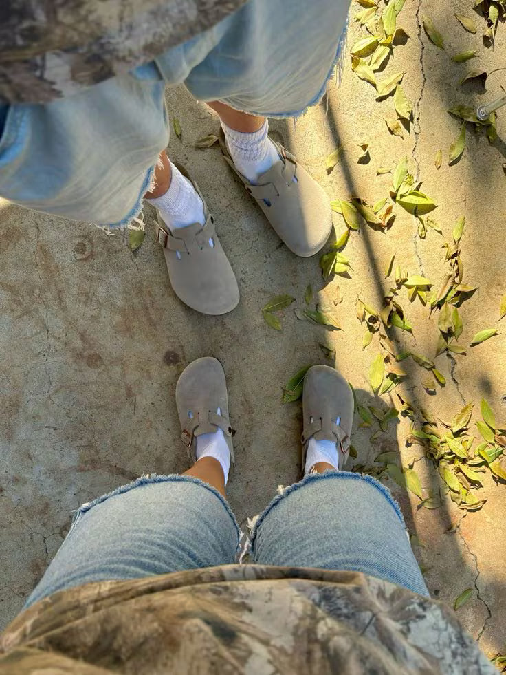 Person wearing beige shoes and light blue jeans standing on a sandy surface with green leaves.