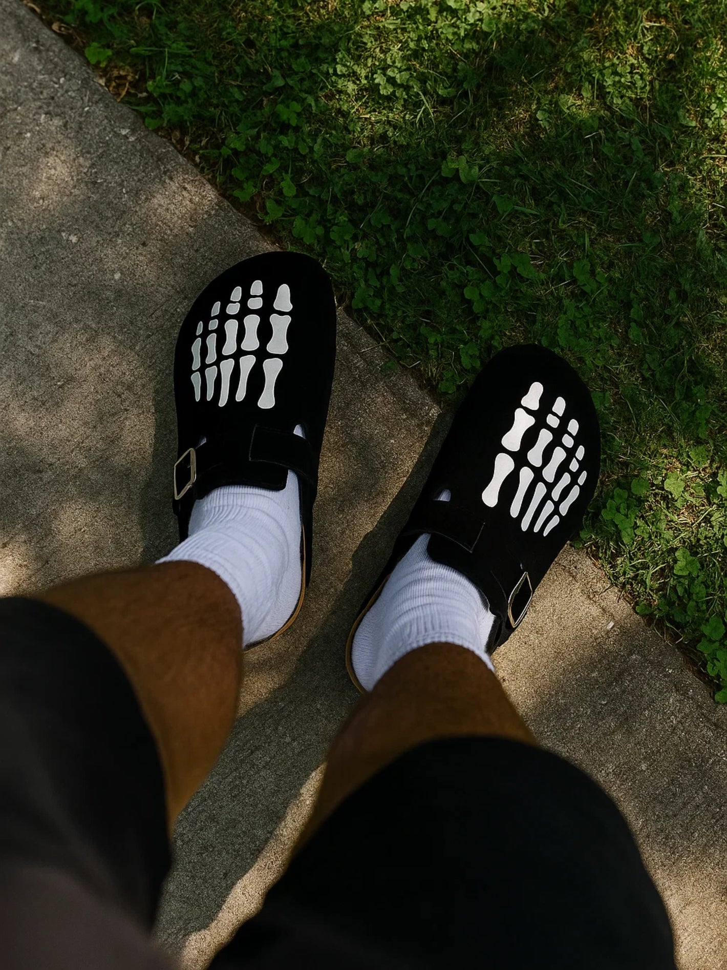 Black sandals with white sole patterns worn on a concrete surface with grass in the background